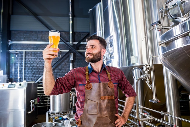 Young brewer wearing a leather apron is testing beer at a modern brewery