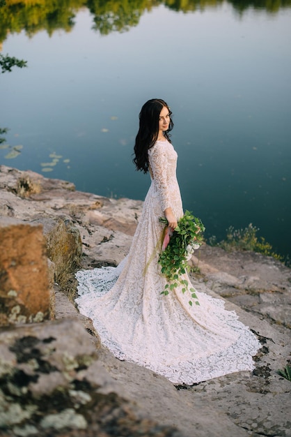 Young bride in wedding dress standing on rocky river bank, sunset