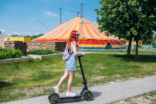 Young bright woman smiles and rides electric scooter in the summer in the city