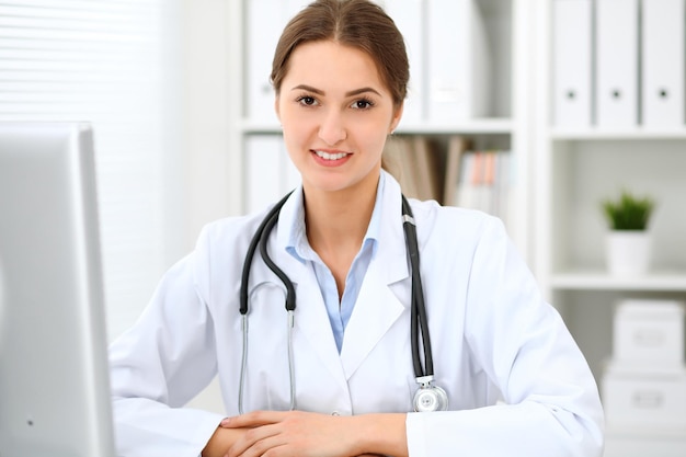 Young brunette female doctor sitting at the table and working at hospital office.  Health care, insurance and help concept. Physician ready to examine patient