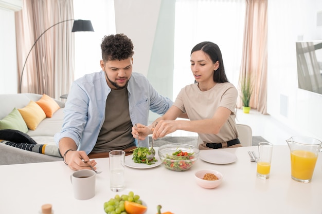 Young brunette female putting fresh vegetable salad on plate of her husband while both going to have breakfast after cooking it