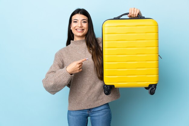 Young brunette girl over blue wall in vacation with travel suitcase