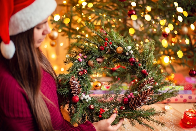 Young brunette girl in christmas hat, holding colorful decorated wreath and looking at it