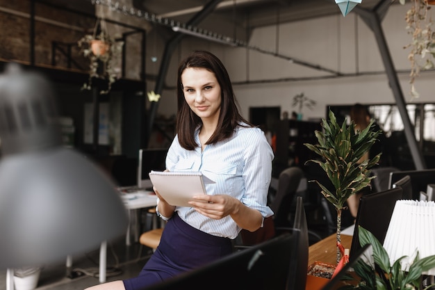 Young brunette girl dressed in office style clothes makes notes in a notebook standing in the modern office