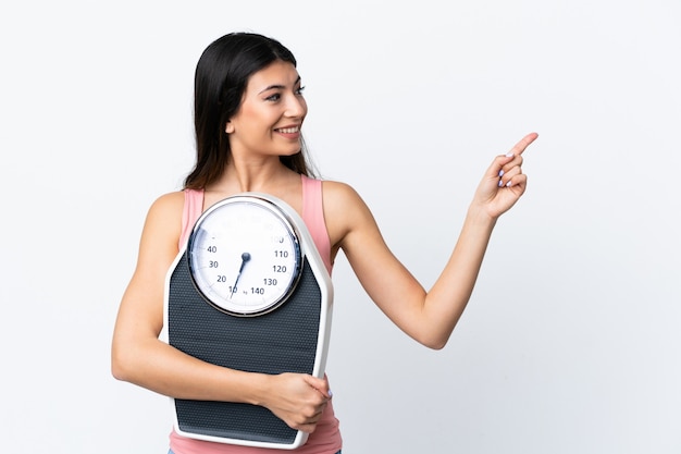 Young brunette girl over isolated white wall with weighing machine and pointing side