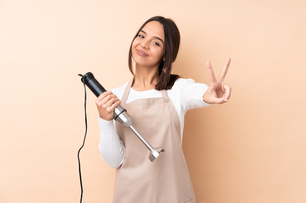 Young brunette girl using hand blender over isolated background smiling and showing victory sign