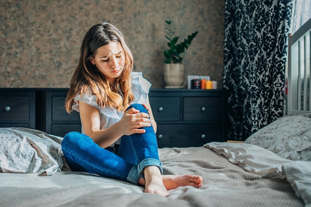 Young brunette girl in a white blouse and blue jeans sits on a bed in her room, clutching a sore knee