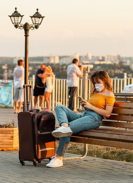 Young brunette girl with a surgical mask browsing through her phone while sitting in park with her suitcase near her.