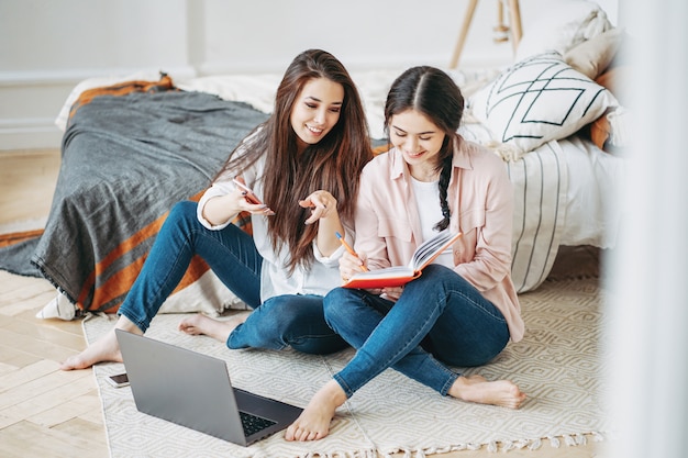 Young brunette girls friends students in casual doing project together looking for information in internet by mobile and laptop at room