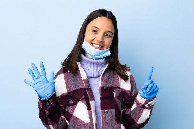 Young brunette mixed race woman protecting from the coronavirus with a mask and gloves over isolated blue wall counting six with fingers