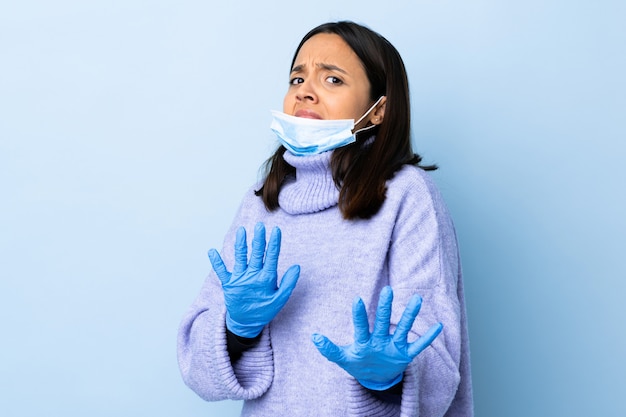 Photo young brunette mixed race woman protecting from the coronavirus with a mask and gloves over isolated blue wall nervous stretching hands to the front