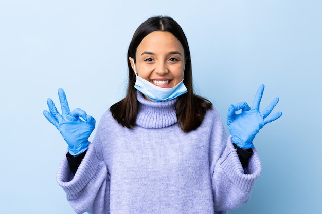 Young brunette mixed race woman protecting from the coronavirus with a mask and gloves over isolated blue wall showing ok sign with two hands