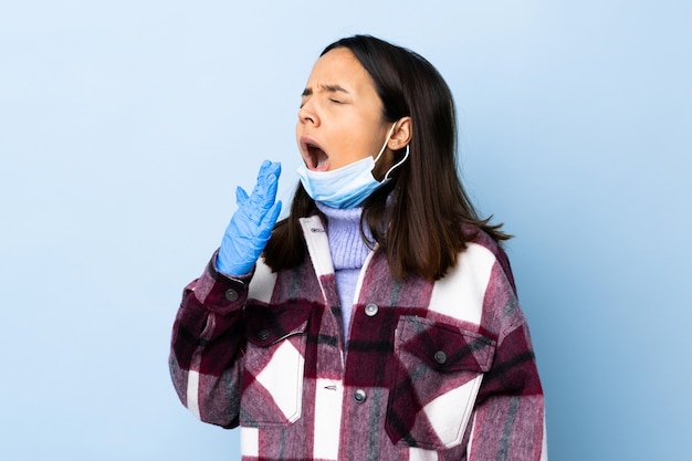 Young brunette mixed race woman protecting from the coronavirus with a mask and gloves over isolated blue wall yawning and covering wide open mouth with hand