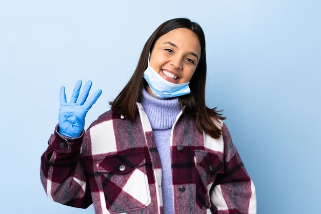 Young brunette mixed race woman protecting with a mask and gloves over blue wall happy and counting four with fingers