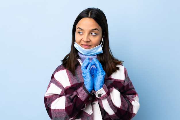 Young brunette mixed race woman protecting with a mask and gloves over blue wall scheming something