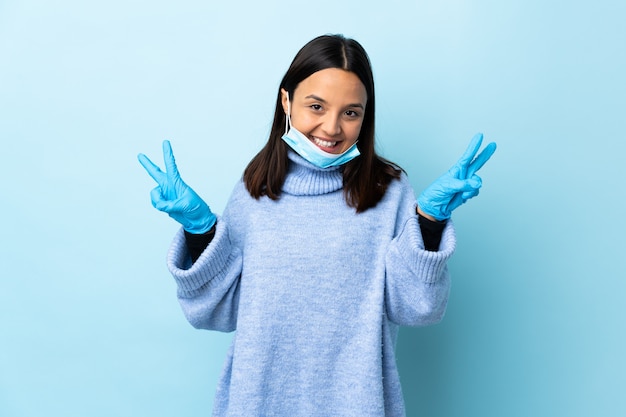 Young brunette mixed race woman protecting with a mask and gloves over blue wall showing victory sign with both hands