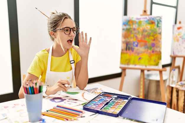 Young brunette teenager at art studio shouting and screaming loud to side with hand on mouth. communication concept.