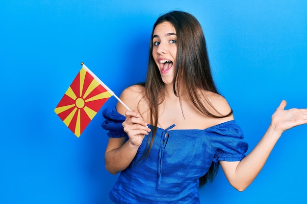 Young brunette teenager holding macedonian flag celebrating achievement with happy smile and winner expression with raised hand