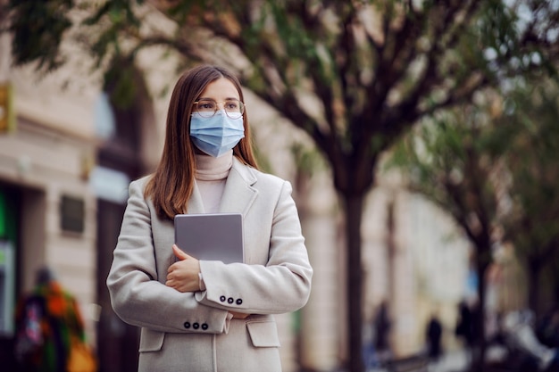 Young brunette with face mask on standing on the street and holding tablet