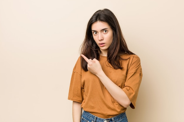 Young brunette woman against a beige background pointing to the side