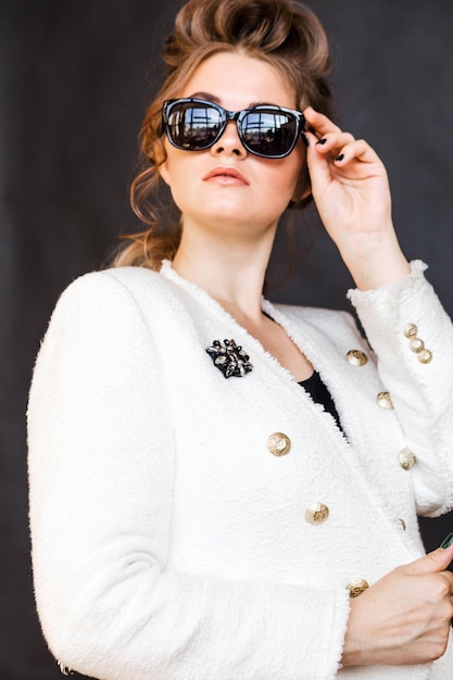 Young brunette woman in black sunglasses and a white jacket with a brooch posing on a black background