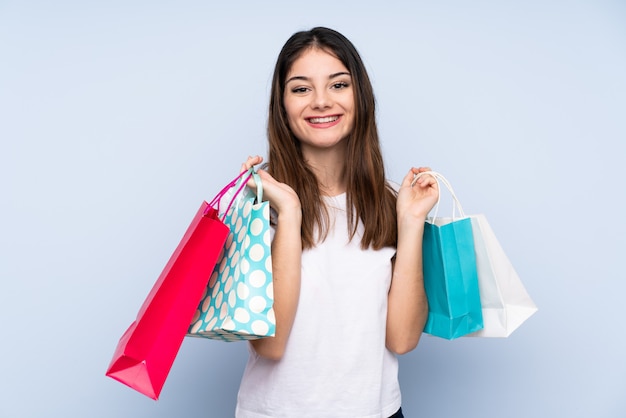 Young brunette woman over blue wall holding shopping bags and smiling