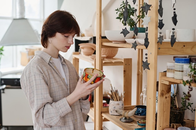 Young brunette woman choosing gift in earthenware shop while holding clay bowl