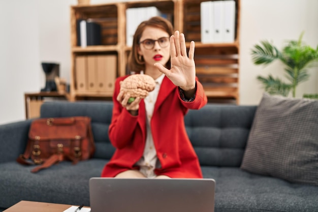 Young brunette woman at consultation office holding brain with open hand doing stop sign with serious and confident expression, defense gesture