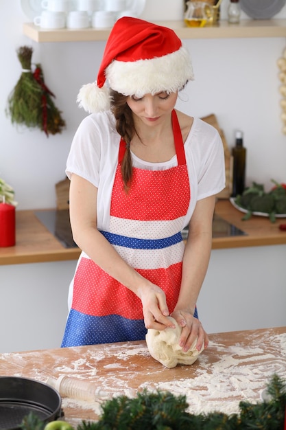 Young brunette woman cooking pizza or handmade pasta while wearing Santa Claus cap in the kitchen. Housewife preparing dough on wooden table. Happy New Year, Marry Christmas, Dieting, food and health