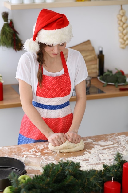 Young brunette woman cooking pizza or handmade pasta while wearing Santa Claus cap in the kitchen. Housewife preparing dough on wooden table. Happy New Year, Marry Christmas, Dieting, food and health