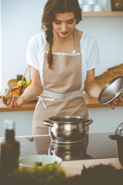 Young brunette woman cooking soup in kitchen. Housewife holding wooden spoon in her hand. Food and health concept.