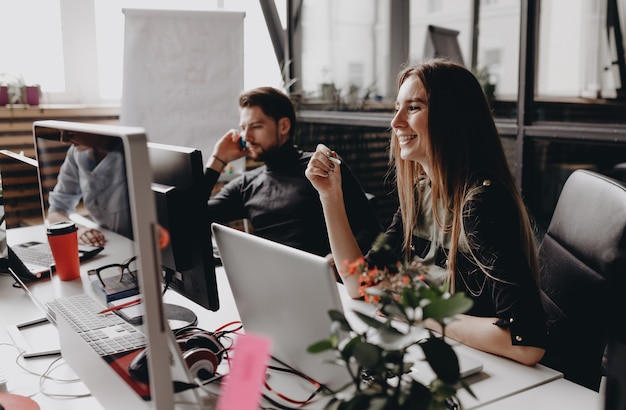 Young brunette woman dressed in office style clothes is working at the computer sitting at the desk with the colleagues in the modern office