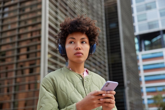 Young brunette woman enjoys music with headphones holds mobile phone downloads song to playlist wears jacket