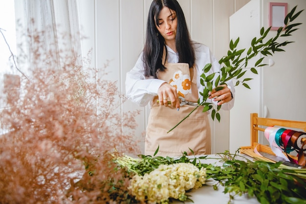 A young brunette woman florist in apron cuts leaves from the stems before starting to create a bouquet in her workshop