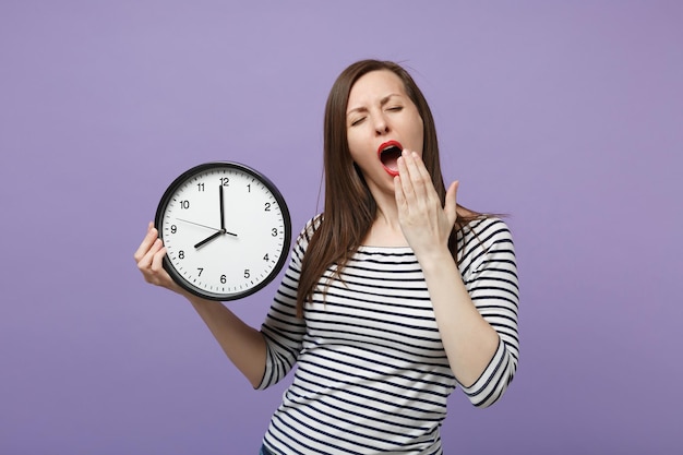 Young brunette woman girl in casual striped clothes posing isolated on violet purple background studio portrait. People sincere emotions lifestyle concept. Mock up copy space. Hold in hand round clock