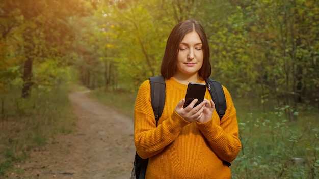 Young brunette woman hiker with backpack in stylish orange sweater texts on mobile phone standing on ground road in green forest closeup