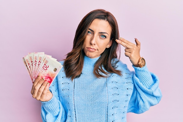 Young brunette woman holding 20 israel shekels banknotes shooting and killing oneself pointing hand and fingers to head like gun, suicide gesture.