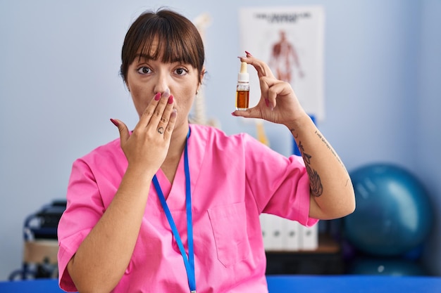 Young brunette woman holding cbd oil at physiotherapy clinic covering mouth with hand, shocked and afraid for mistake. surprised expression