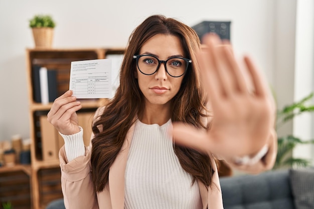 Young brunette woman holding covid record card with open hand doing stop sign with serious and confident expression, defense gesture