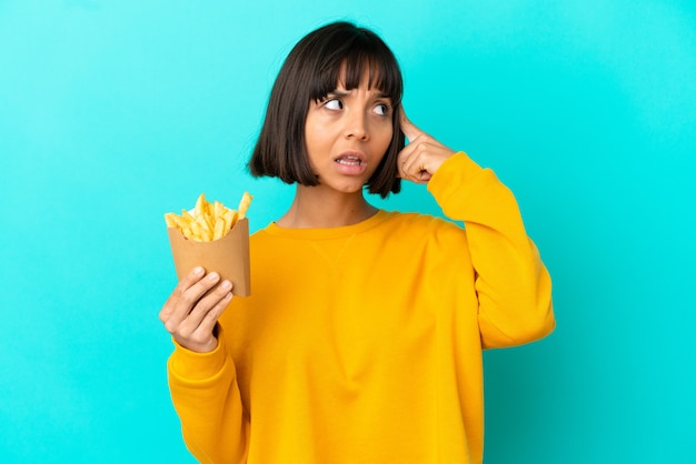 Young brunette woman holding fried chips over isolated blue background having doubts and thinking