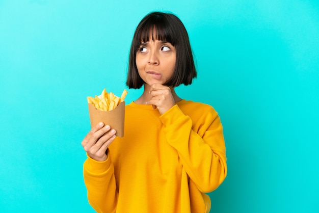 Young brunette woman holding fried chips over isolated blue background having doubts and thinking