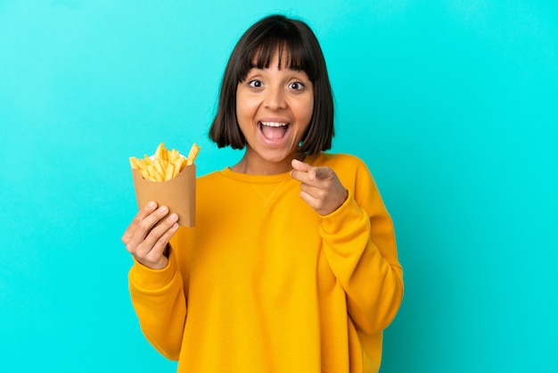 Young brunette woman holding fried chips over isolated blue background surprised and pointing front