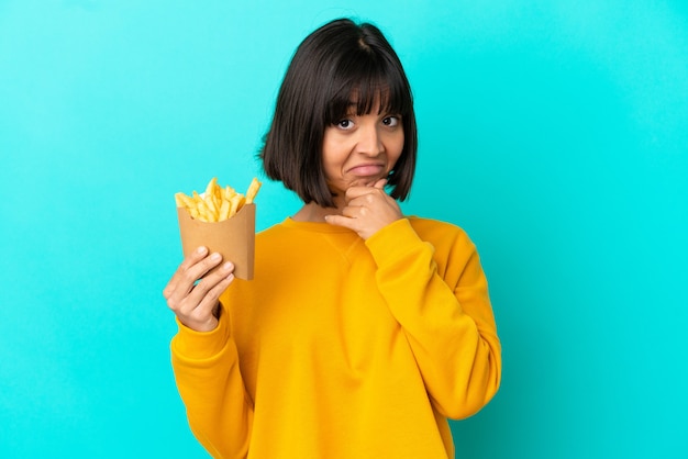 Young brunette woman holding fried chips over isolated blue background thinking