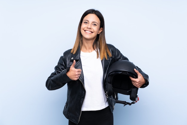 Young brunette woman holding a motorcycle helmet over blue wall with thumbs up