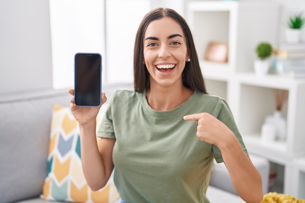 Young brunette woman holding smartphone showing blank screen pointing finger to one self smiling happy and proud