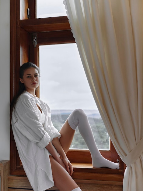 Young brunette woman at home in the morning Sensual young woman with sexy body in shirt and white lingerie in modern apartment near window after waking up weekend vacation