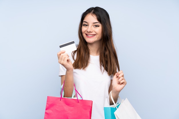 Young brunette woman over isolated blue wall holding shopping bags and a credit card