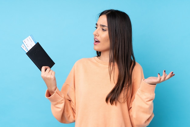 Young brunette woman over isolated blue wall in vacation with plane tickets and surprised