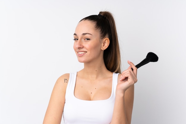 Young brunette woman over isolated white wall holding makeup brush and looking up