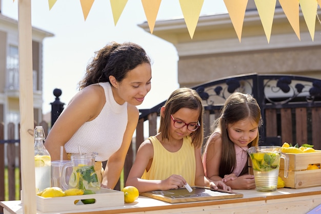 Young brunette woman looking at her daughter pointing at notice on small board with piece of chalk while selling lemonade on sunny day